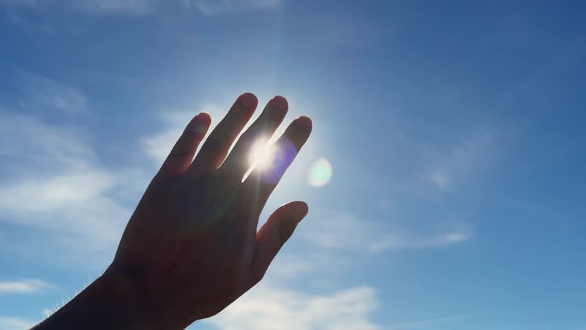 Close-up hand in sunshine outdoors, stretching arm to sun rays on blue sky with white clouds. Nature concept.