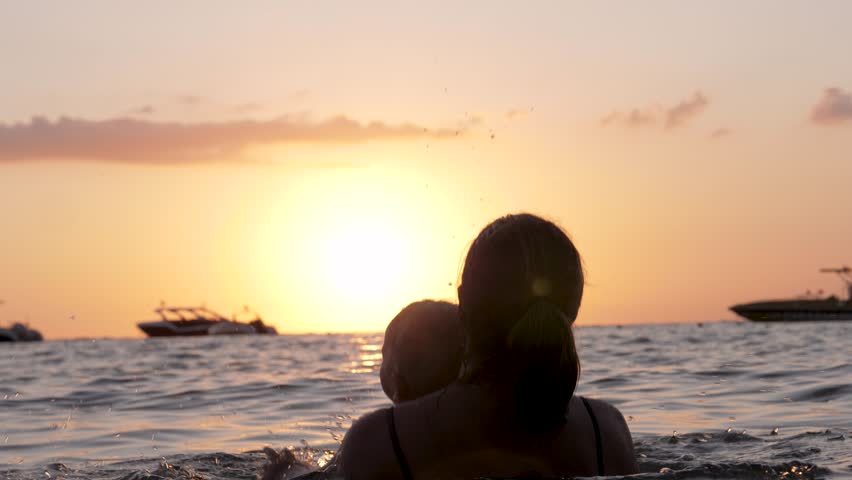 Backlit silhouette mother and baby swimming funny during golden sunset, sharing tender moment of connection against vibrant orange yellow sky and ocean waters. Mom throws her baby up against sunset