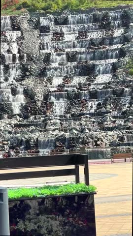 Water flows gracefully down man made waterfall, creating soothing ambiance in city park setting, with planter and bench in foreground. waterfall