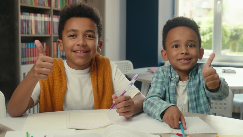 Happy little boys African American kids students pupils schoolboys friends siblings smiling children thumbs up gesture school primary education studying together writing desk classroom books learning