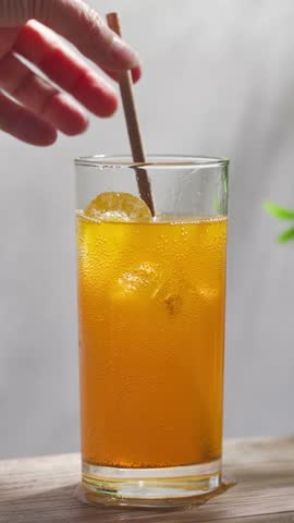 hand put straw in fresh sparkling orange juice drink in glass on sunny wooden kitchen tabletop and gray wall background, slow motion