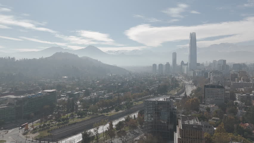 Backwards drone shot of Santiago de Chile in South America watching over the skyscrapers and big buildings with snowy mountains in the background on bright but cloudy day LOG