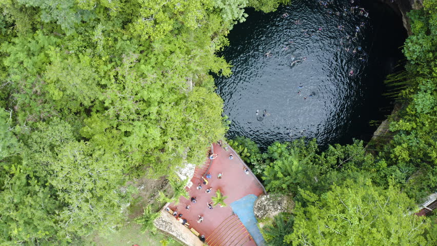 An aerial view of a cenote hidden within lush jungle, with a rustic building nearby. The deep blue water contrasts beautifully with the surrounding green foliage.