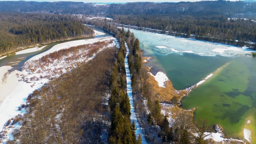 Aerial view of a snow-covered path lined with evergreen trees between a partially frozen river and a green water body in a winter landscape
