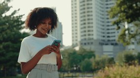 Portrait of happy woman sending message on smartphone in park against skyscrapers. African American female smiles looking at mobile phone in urban environment. City lifestyle - Powered by Shutterstock - Get 15% off with code: PIKWIZARD15