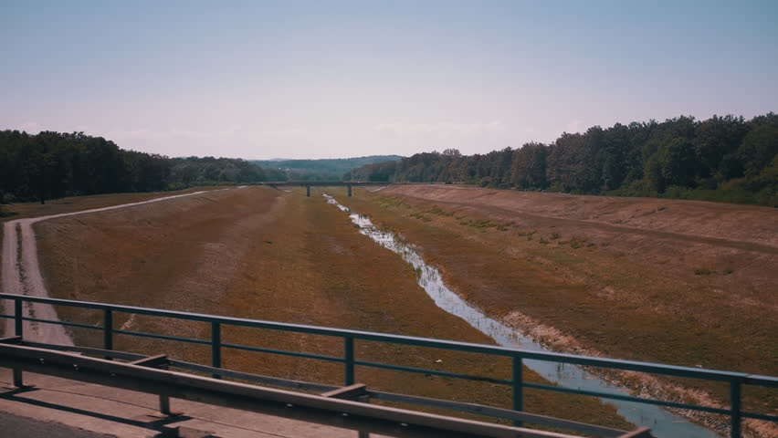 Color-corrected view of a dried riverbed from a moving car, highlighting the dry landscape, trees, and a distant bridge. This footage represents climate change and environmental challenges.