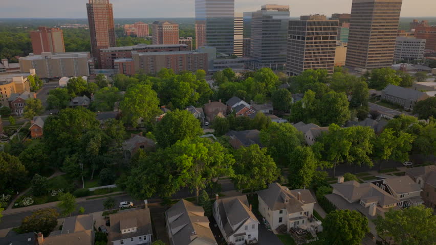 Aerial shot over Clayton neighborhood houses, tilting up to reveal city office buildings at sunset in St. Louis, Missouri. Warm golden hues highlight the suburban charm and urban skyline