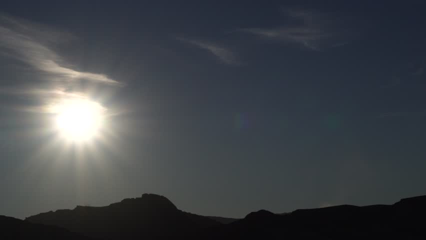 A blazing and dazzling sun in the sky under a sky that resembles a work of art, all with the hilltop of a desert environment.