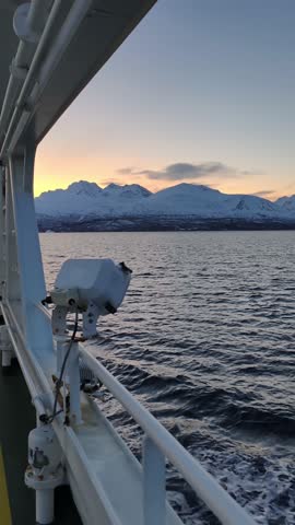 A stunning Arctic landscape is framed by the metal structure of a ferry, creating a natural window to the snow-capped mountains beyond. The icy waters stretch toward the rugged peaks