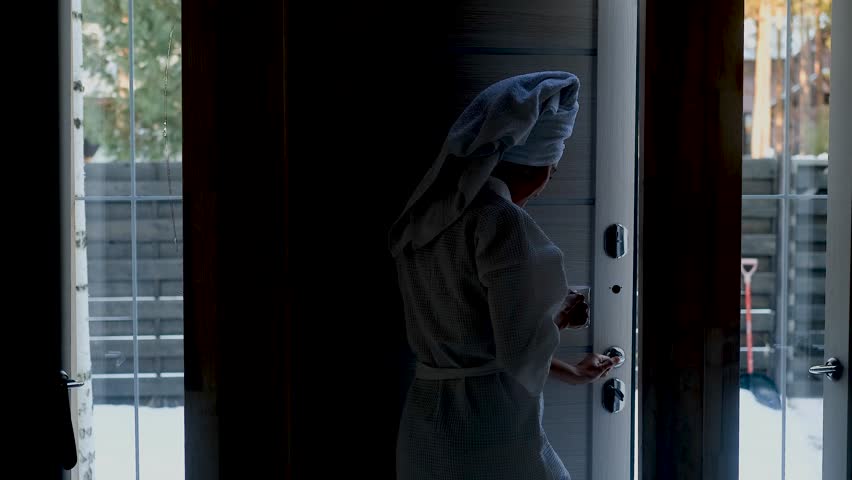 A young woman in a bathrobe and a towel on her head holds a cup of herbal tea in her hands. She came out of the bathhouse into the fresh cold air on a winter day