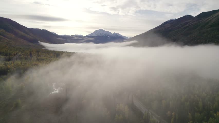 Mystic fog and clouds over national park with forest trees and snowy mountains in background. Aerial revealing wide shot. Sunrise in Alaska, USA.