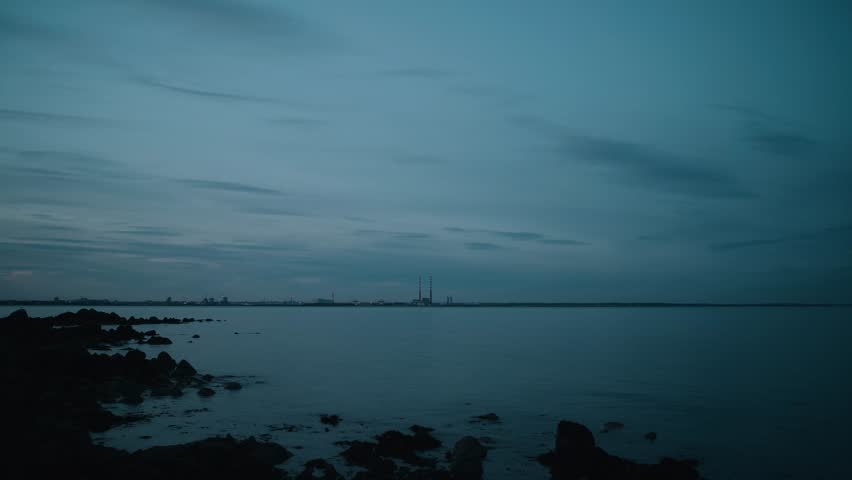 A locked-off shot of Poolbeg Towers in Dublin, Ireland, seen across calm coastal waters at dusk. The industrial skyline contrasts with the darkening sky, creating a moody and atmospheric scene.