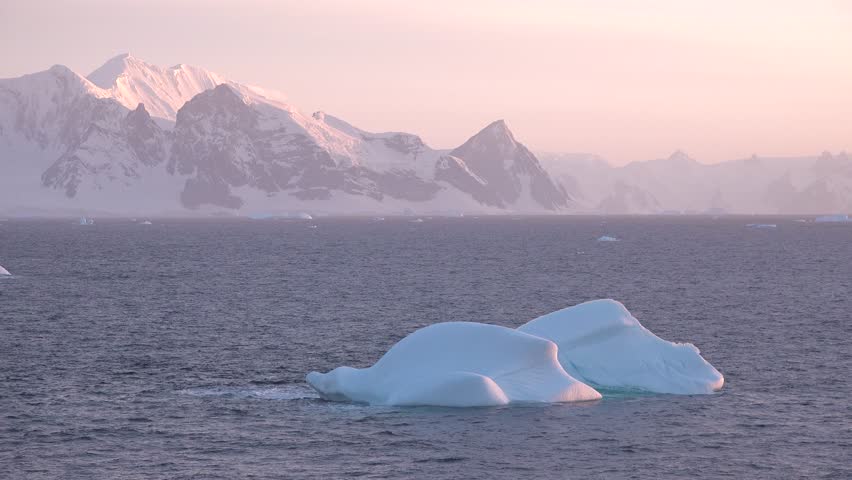 The sky is painted with colors as the sun sets behind a large iceberg floating serenely in the Arctic Ocean, surrounded by distant ice formations and a calm atmosphere.