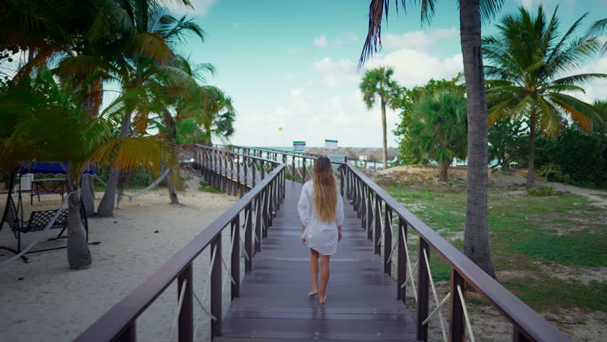 Aerial follow shot with happy woman vacationer walking in tropical garden. Summer in south, beautiful lady resting and enjoying life in hotel with big territory and path to ocean, trip to Caribbean