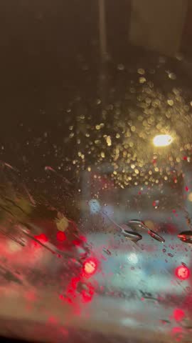 A closeup of raindrops falling gently and softly on the darkened glass surface with blurry lights
