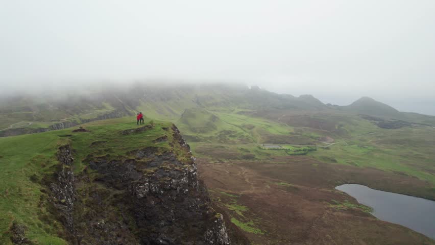 Epic Wide Aerial Adventure Couple on Cliff Orange Coats Quiraing Scotland Isle of Skye Fog Scottish Highlands Landscape Drone Over and Flies Far Away