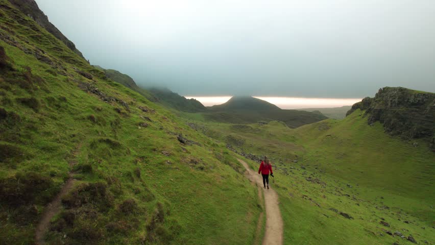 Epic Drone Follows Female Photographer Hiking Remote Trail in Scottish Highlands. Orange Coat Girl Walks the Quiraing Cloudy Morning Aerial