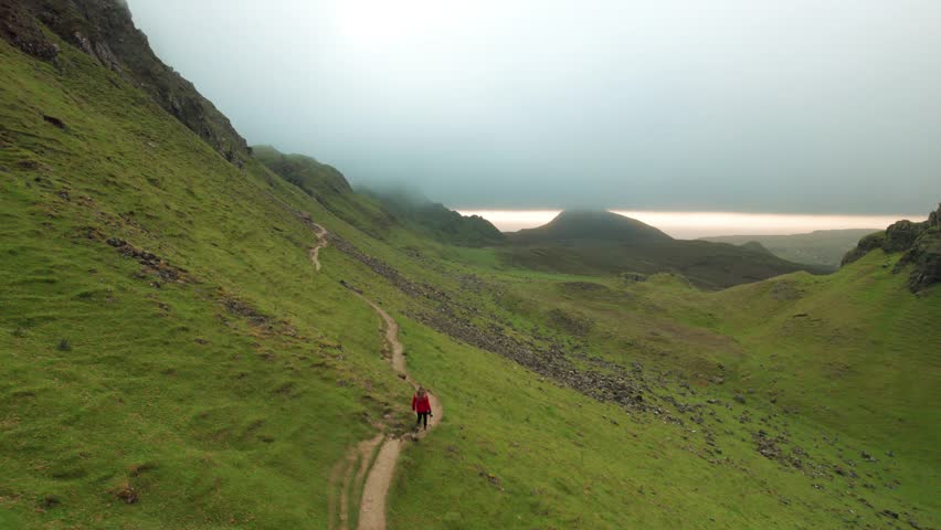 Epic Drone Follows Female Photographer Hiking Remote Trail in Scottish Highlands. Orange Coat Girl Walks the Quiraing Cloudy Morning Aerial