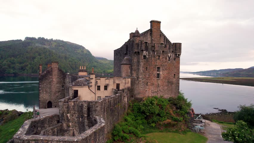 Aerial Flight From Eilean Donan Castle Scotland Backwards Along Stone Bridge. Morning Cloudy Landscape.