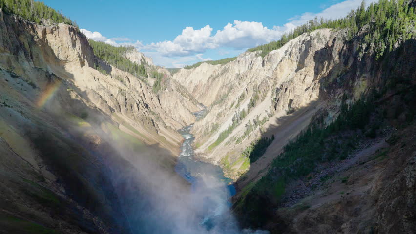 Mist rises from a powerful waterfall in Yellowstone, forming a stunning rainbow over the river valley, enhancing the park’s natural beauty.