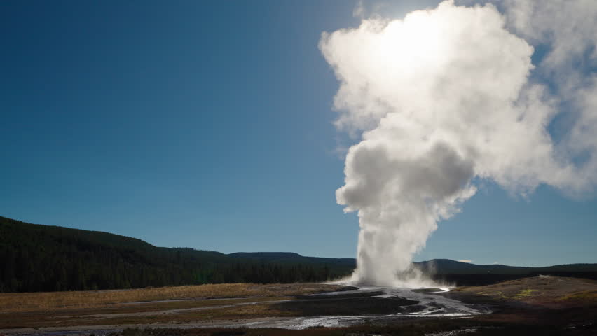 Rays of sunlight pierce through the steam of a Yellowstone geyser, casting a golden glow over the forest trees, beneath clear blue skies.