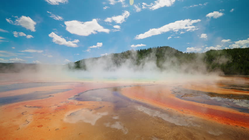 The stunning Grand Prismatic Spring in Yellowstone steams under a bright summer sky, showcasing its vibrant colors and unique geothermal beauty.