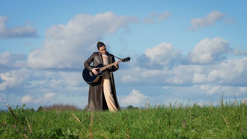 Female guitarist performing acoustic melody amid lush grassy landscape, wearing long coat and standing against expansive blue sky with soft clouds, embodying musical creativity