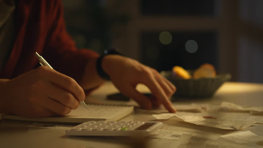 A person is seated at a table, taking notes in a notebook while reviewing bills and using a calculator. The warm lighting creates an evening atmosphere, suggesting they are managing personal finances