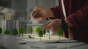 A person is designing a digital urban model by sketching on a tablet with a stylus, surrounded by miniature buildings and trees. This activity takes place in a modern design studio - Powered by Shutterstock - Get 15% off with code: PIKWIZARD15
