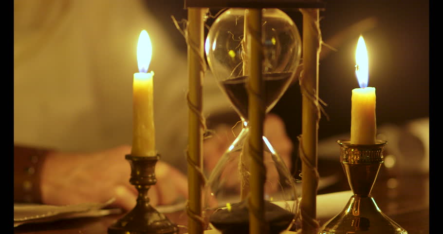 A scene from the dark ages or medieval era of a man working at a desk with an hourglass in the foreground and focus on a candle.