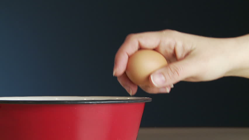 Hand cracks a fresh egg on the side of a red metal bowl and drops it into the