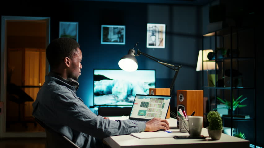 Financial manager doing remote accounting work with music on headset, solving his tasks on laptop at home. African american guy multitasking with his favorite playlist to help him focus. Camera A.