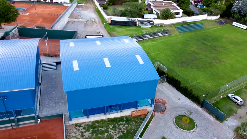 An aerial orbit drone shot of the newly completed sports complex in Quito, Ecuador. The image shows the blue-roofed buildings, tennis courts, and the surrounding green fields.