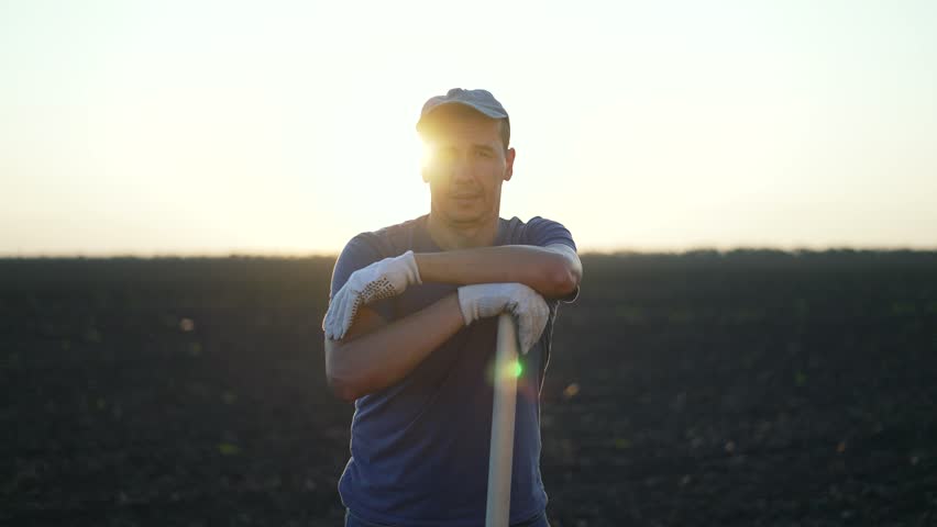 Male farmer with shovel rural field. Portrait of farmer resting on shovel at sunset. Farmer in gloves posing in rural landscape. Hard work in farming concept. Farmer portrait with shovel rural setting