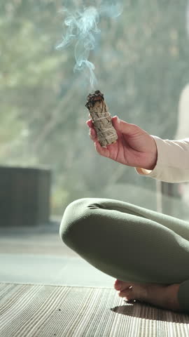 Wellness practitioner performing sage smudging ritual, waving burning white sage bundle while meditating, cleansing negative energy and promoting inner peace and tranquility