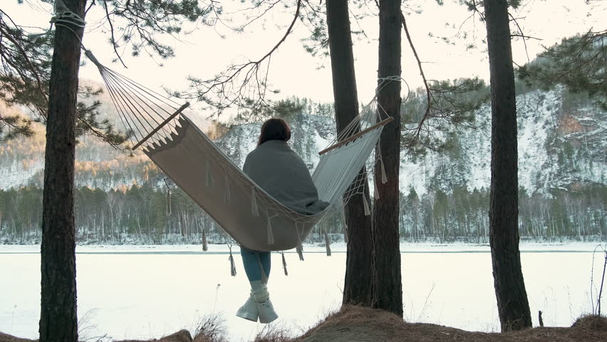 Winter landscape featuring young woman relaxing in hammock strung between pine trees, overlooking frozen lake during serene sunset with snow-covered surroundings