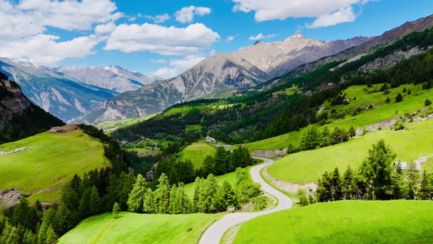 A drone breathtaking view of a lush green valley with winding road and remote house on top of a hill under clear skies in Col de la Bonette, France