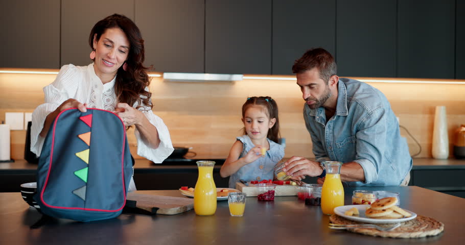 Happy family, child or morning with bag for school, breakfast or getting ready together in home kitchen. Mom, dad or daughter with lunch, meal or food in preparation for kindergarten snack at house