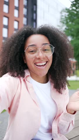 POV Vertical video African american female student talking on video call using phone standing in campus near university building. Happy black woman greeting, smiling, speaking online looking at camera