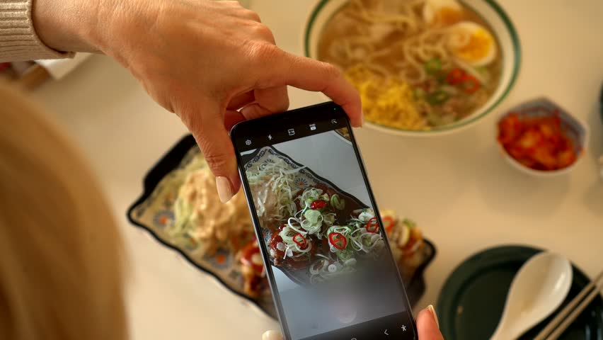 Food blogger captures meal before eating. Close up of woman's hands holding smartphone, taking photo of colorful Asian dish. Social media trend, culinary photography, restaurant experience.