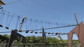 A young, focused woman boldly walks along a swinging suspended walkway in protective gear. A woman walks along the path of a rope park high above the ground. - Powered by Shutterstock - Get 15% off with code: PIKWIZARD15
