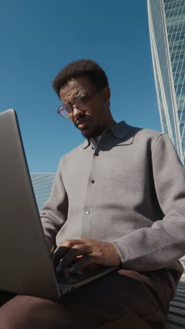 Vertical shot of bearded African American man with short curly hair intently working on his laptop outdoors against modern glass building and clear blue sky
