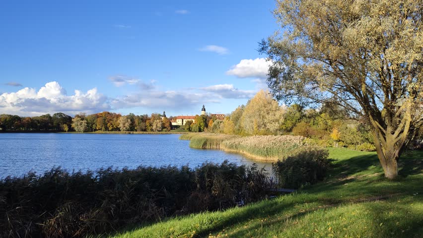 In autumn, the evening sun sinks to the horizon and illuminates the medieval castle and the trees with yellow and red leaves standing on the grassy banks. Reeds grow in the water. Blue sky with clouds