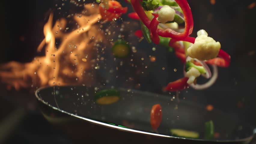 Closeup of chef preparing and throwing vegetable mix on frying pan on fire. Preparation fresh appetizing food. Vegetables flying into the pan in slow motion. Healthy vegetarian food cooking close-up
