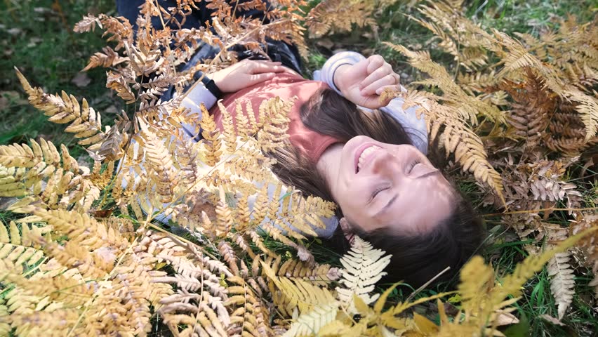 Cheerful young woman lies on yellow autumn leaves in the park and smiles.