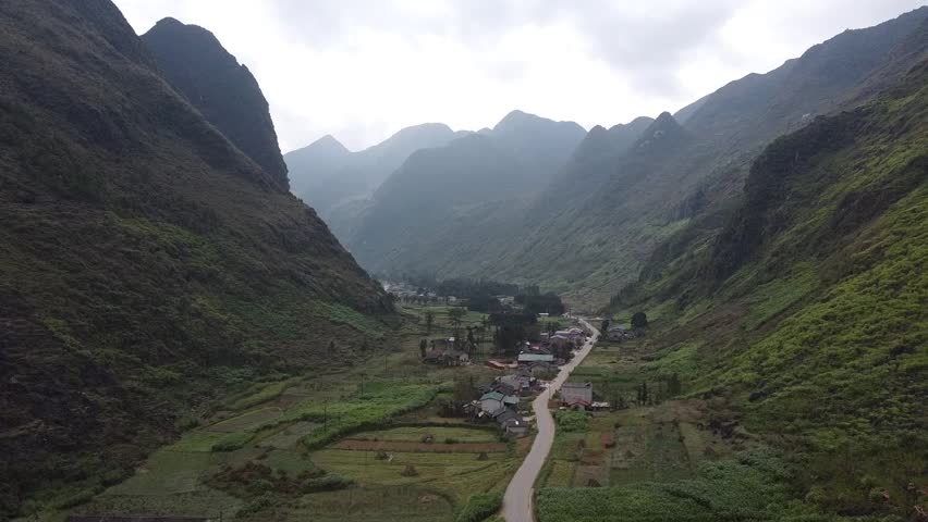 Remote valley in Vietnam with a winding road leading to a small village. Towering mountains surround green fields and scattered houses. Misty peaks rise in the background under a cloudy sky.