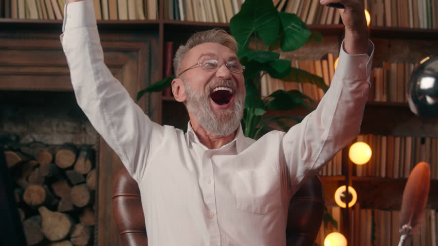 Caucasian old man mobile phone smartphone victory triumph businessman business excited happy elderly male winner home office indoor mature senior online betting gambling success lottery achieve joyful