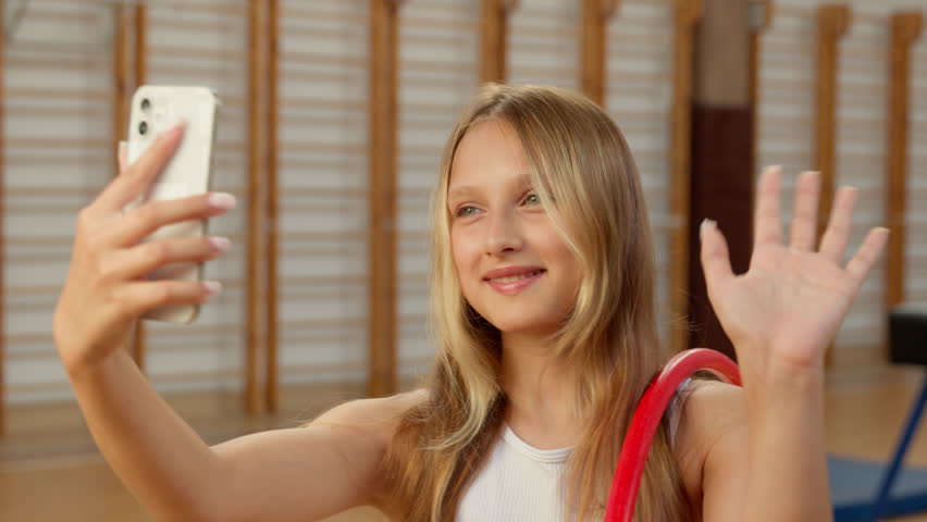Caucasian little girl holding mobile phone smartphone blogger influencer video call online child kid schoolgirl school sport gym waving hand greeting vlog daughter app conference gymnast hoop student