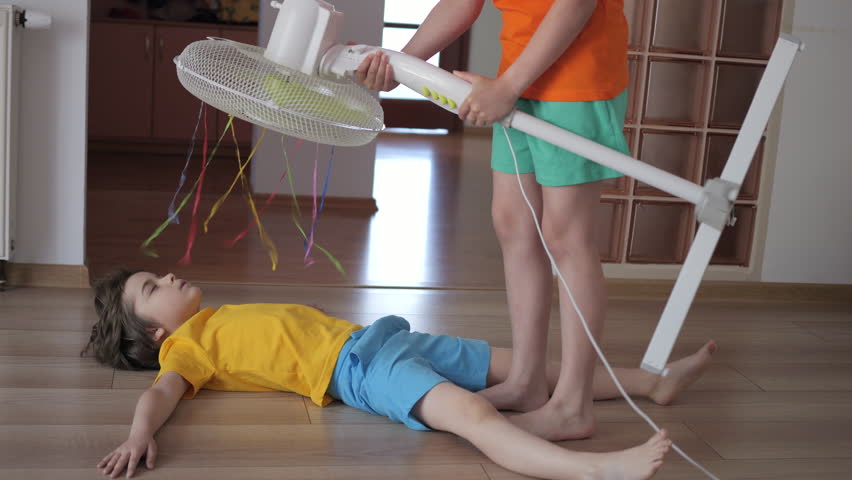 Boy and Girl in Front Fan Hot Day on Home in Living Room. Kids Enjoying Cool Wind From Electric Fan at Home Summer Heat Weather. Children Suffering From Summer Heat Enjoy Fresh Cool Breeze Blowing Fan