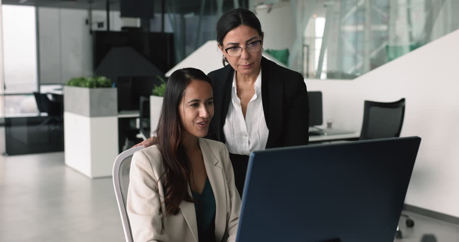 Successful young employee woman showing completed task on monitor to mentor. Elder business professional woman, boss, manager giving support to new worker, touching shoulder, training intern